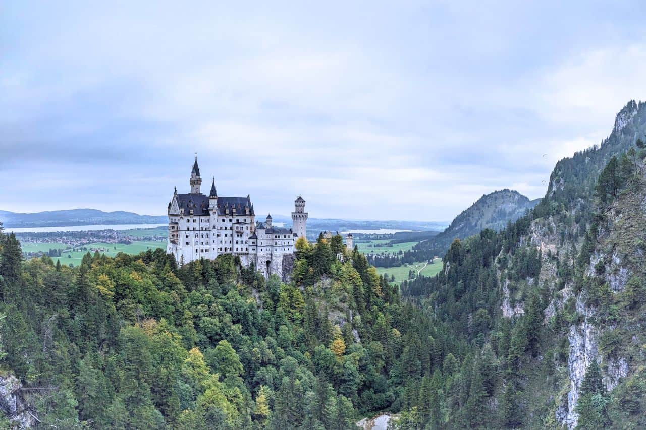 Neuschwanstein Castle in the Bavarian Alps under a blue sky, a white neo-Romanesque castle with turrets and spires, surrounded by green trees.