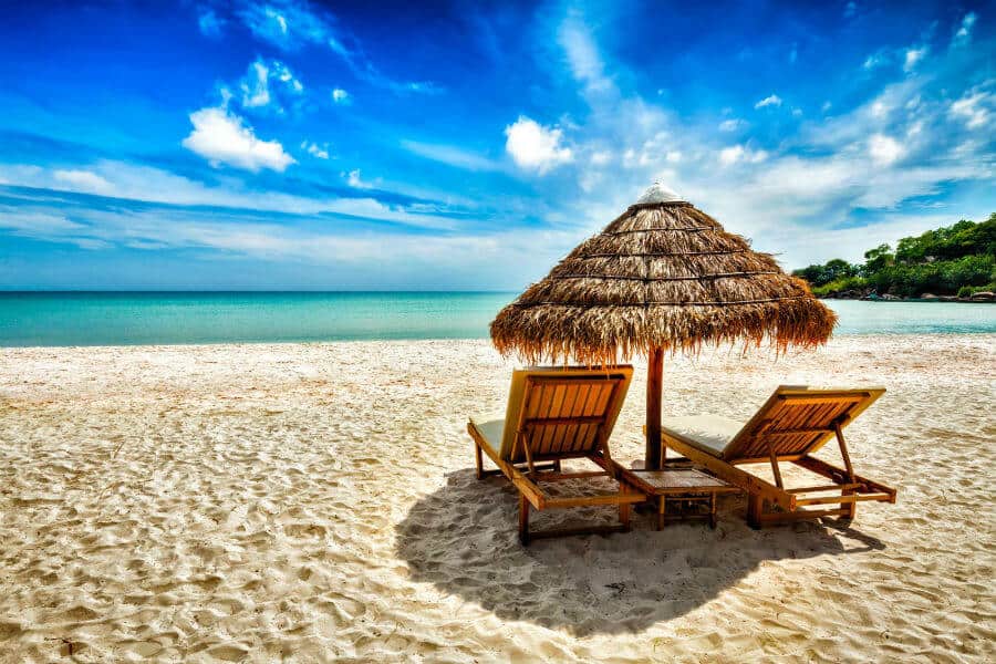 Two wooden chairs with a wicker umbrella on white sand and blue skies as one of the beaches in southeast asia