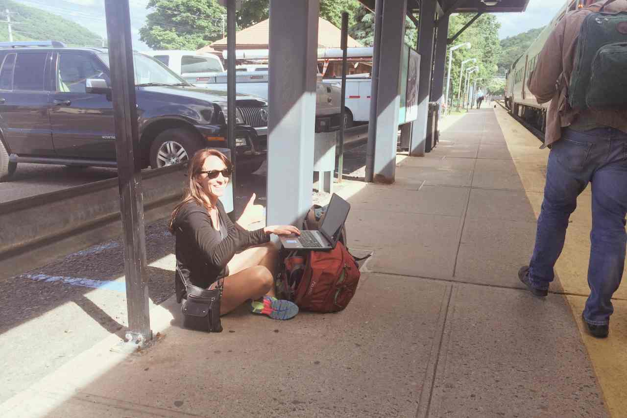 woman sitting on a train platform using her red ladies backpack for travel as a desk 