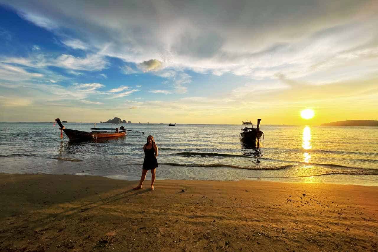 woman standing on a thailand beach with two boats and a sunset