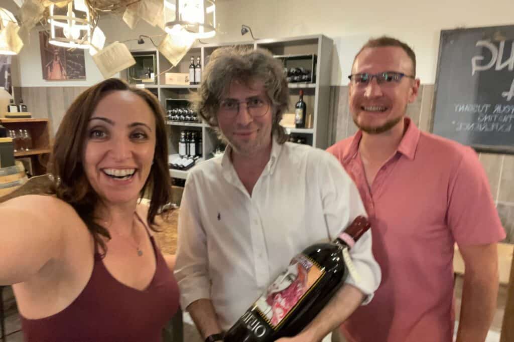 three people inside of a winery smiling holding a large bottle of red wine