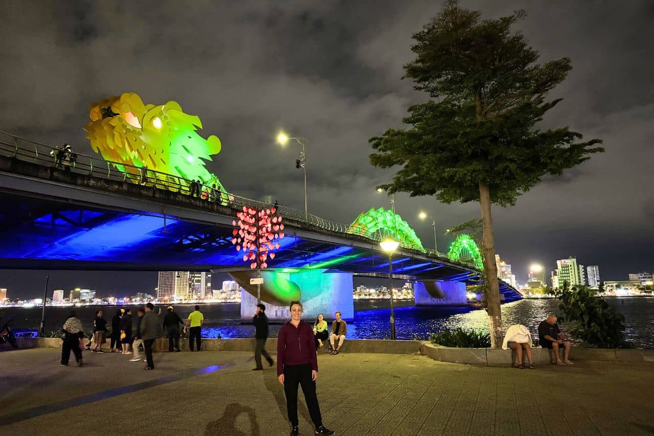 The iconic Dragon Bridge in Da Nang glowing in green and blue lights with people enjoying riverside views